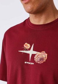 Primer plano de una persona que lleva una camisa burdeos con una estrella blanca y dos flores naranjas bordadas sobre el logotipo "PXP" en el pecho.