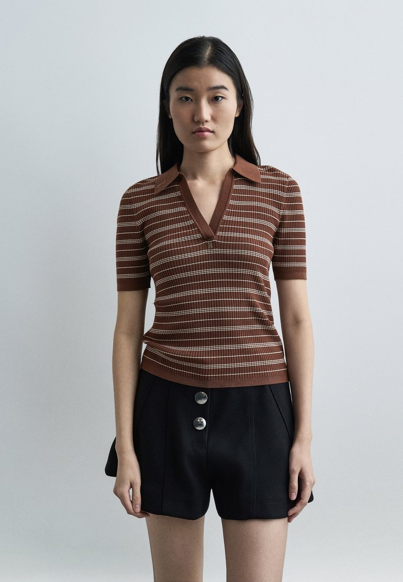 Jeune femme portant un haut à manches courtes à rayures marron avec col et un short taille haute noir avec deux grands boutons, debout devant un fond uni.