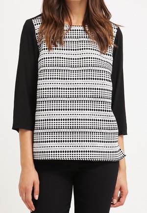 Kvinde iført en sort og hvid mønstret ærmeløs top over en sort langærmet skjorte, kombineret med sorte bukser.