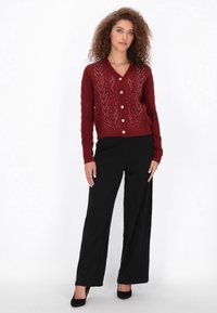 Burgundi színű kötött kardigán V-nyakkal és gyöngygombokkal, kábelmintákkal és díszítésekkel, fekete, bő szárú nadrággal párosítva.