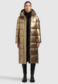 Giacca imbottita color oro con cappuccio, caratterizzata da un design lungo, motivo trapuntato e chiusura con cerniera. Indossata sopra un outfit nero, abbinata a stivali al ginocchio.