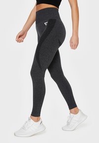 Leggings negros de cintura alta con un acabado texturizado, que presentan un diseño de costura lateral y un pequeño acento de logo, combinados con zapatillas deportivas blancas.