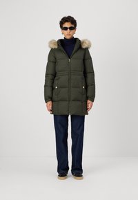 Zaļš polsterēts mētelis ar kažokādas oderi kapucei, ar rāvējslēdzēja aizdari, sānu kabatām un zeltainiem aksesuāriem, apvienots ar tumšām biksēm un melniem apaviem.