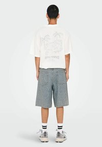 Camiseta blanca oversize con diseño de palmera y texto en la parte posterior, combinada con pantalones cortos azules con estampado, calcetas blancas y zapatillas grises.