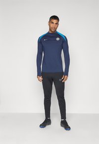Maglietta sportiva a maniche lunghe blu navy con accenti blu, dotata di colletto con zip e logo della squadra, abbinata a pantaloni neri aderenti e scarpe nere.