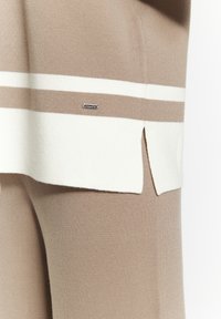 Neulepaita beige-värissä, jossa on valkoisia raitoja. Sisältää sivu-reiän ja hienovaraisen logotagin. Kangas näyttää pehmeältä ja teksturoidulta.