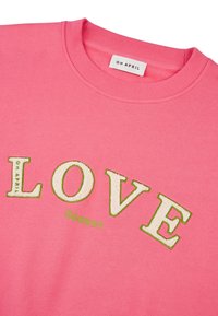 Růžová mikina s žebrovaným výstřihem, na které je nápis „LOVE“ vyšitý bílou a zelenou texturou a pod ním menší zelený nápis „Season“.