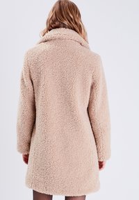 Beige, flauschiger Mantel aus einem strukturierten, schaffellig ähnlichem Material, mit einem breiten Kragen und langen Ärmeln, der gerade über das Knie fällt.