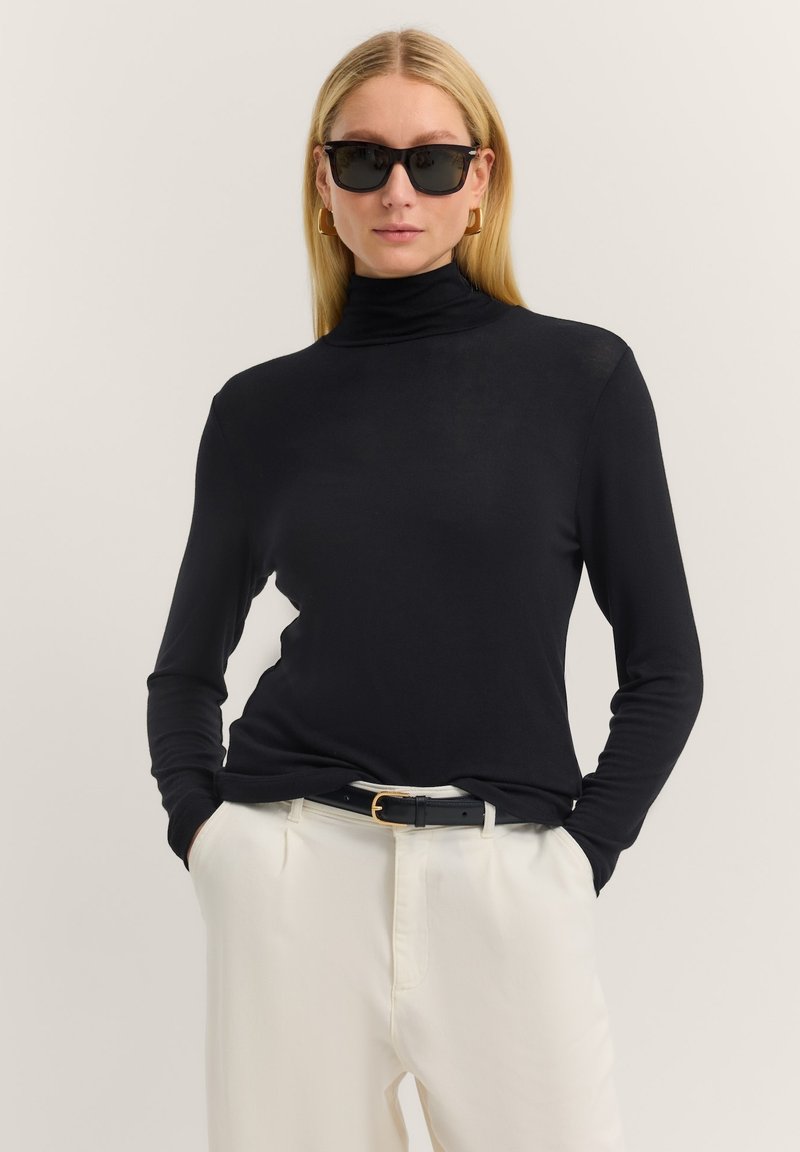 Schwarzes langes Turtleneck-Oberteil aus glattem Stoff, kombiniert mit cremefarbenen Hosen und einem schwarzen Gürtel mit goldener Schnalle. Trägt schwarze Sonnenbrillen.