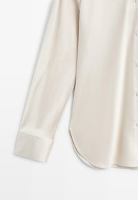 Chemise beige à manches longues avec boutons, ourlet arrondi et poignets larges, présentée sur un fond blanc.