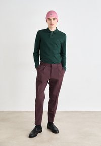 Cămașă polo verde cu mânecă lungă, pantaloni burgundy croiți, căciulă tricotată roz și mocasini negri cu detalii din piele texturată.