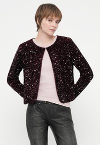 Jaunas žmogus su trumpais rudos spalvos plaukais dėvi purpurinį blizgučių švarką ant šviesiai rožinės spalvos viršaus ir tekstūruotų juodų kelnių, stovėdamas priešais baltą foną.