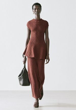 Le mannequin défile avec une tunique texturée couleur rouille et un pantalon large, portant un sac à main noir, sur un fond clair uni.