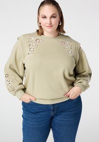 Lichtgroene pullover trui met bloemen uitsnijdingen op de schouders en mouwen. Geribbelde manchetten en zoom. Gecombineerd met een blauwe spijkerbroek.