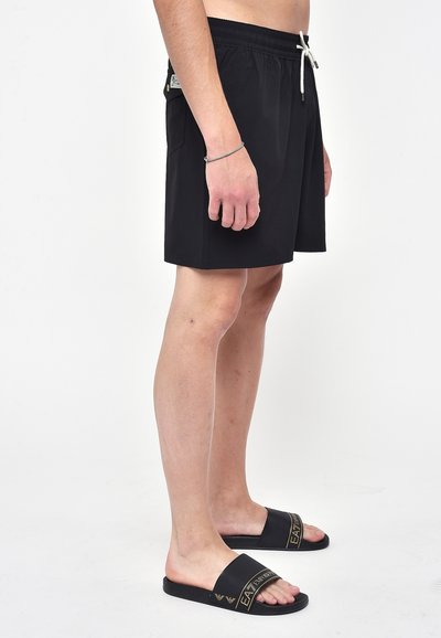 Pantaloni da bagno neri con vita elastica e lacci, dotati di una tasca laterale e un tessuto liscio e leggero. Abbinati a sandali neri scivolosi.