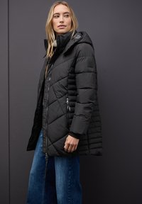 Černá prošívaná péřová bunda s kapucí, která má přední zip a boční kapsy. Texturovaný vzor na rukávech, v kombinaci s modrými džíny.