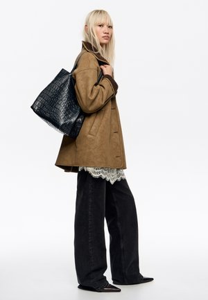 Mujer con cabello rubio lleva chaqueta marrón de borrego, vaqueros negros de pierna ancha, zapatos negros puntiagudos, top blanco de encaje y lleva un bolso grande negro con textura de cocodrilo.