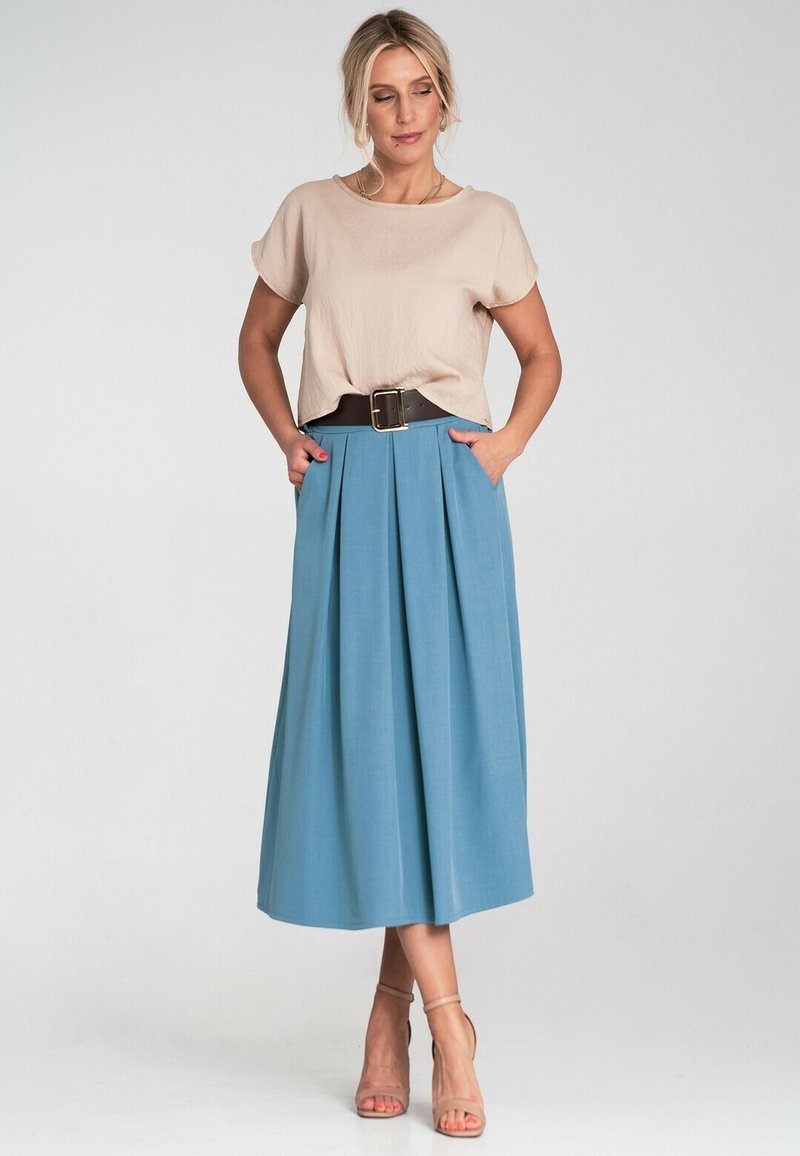 Kobieta w beżowej bluzce z krótkim rękawem, wpuszczonej w niebieską plisowaną midi spódnicę z szerokim czarnym paskiem i cielistych sandałach na kostkę.