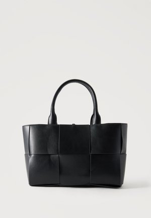 Borsa tote in pelle nera con grandi pannelli quadrati intrecciati e due manici arrotondati su uno sfondo chiaro e semplice.