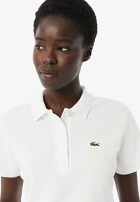 Witte polo-shirt van piqué stof, met een klassieke kraag, een drieknopen sluiting en een klein groen krokodillenlogo op de borst.