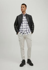 Zwarte leren bomberjack, wit-blauw geruit overhemd, lichtbeige cargobroek en zwarte sneakers. Casual outfit met praktische zakken.