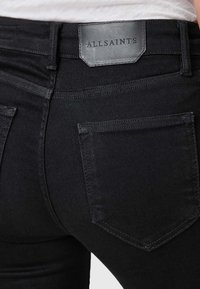 Jean en denim noir à coupe ajustée, avec un patch de marque en cuir visible, deux poches arrière, un tissu texturé et des détails de couture subtils.