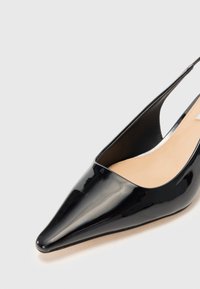 Mustaa lakattua nahkaa oleva slingback-korkokenkä, jossa on terävä kärki, sileä pinta ja minimalistinen muotoilu. Paljastettu kanta-alue ja beige pohjallinen.