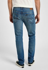 Vaqueros de mezclilla azul con un ajuste slim, cinco bolsillos y un parche trasero de cuero marrón. La tela tiene una textura suave con un ligero desvanecimiento.