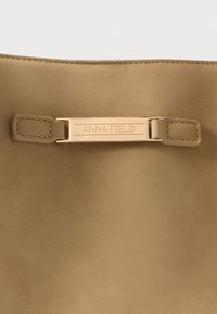 Tanfarvet taske i kunstlæder med en glat tekstur, der har en guldfarvet metalplade med indgraveret "ANNA FIELD" og to fastgørelsestapper.