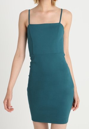 Robe ajustée sans manches couleur sarcelle avec fines bretelles et un décolleté carré portée par une personne devant un fond uni.