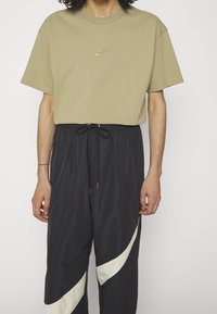 Khaki t-shirt med korta ärmar och broderad Nike-logotyp; kombinerad med svarta träningsbyxor med ett vitt böjt mönster och justerbar snörning.