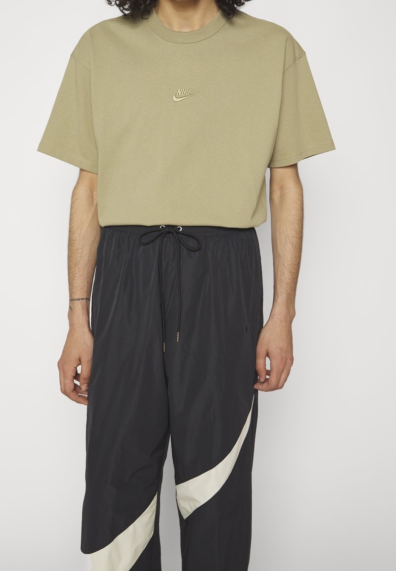 Khaki t-shirt med korta ärmar och broderad Nike-logotyp; kombinerad med svarta träningsbyxor med ett vitt böjt mönster och justerbar snörning.