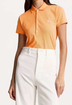 Kvinde iført en orange poloshirt med korte ærmer og logo samt højtaljede hvidebukser, vist fra hals til hofter.