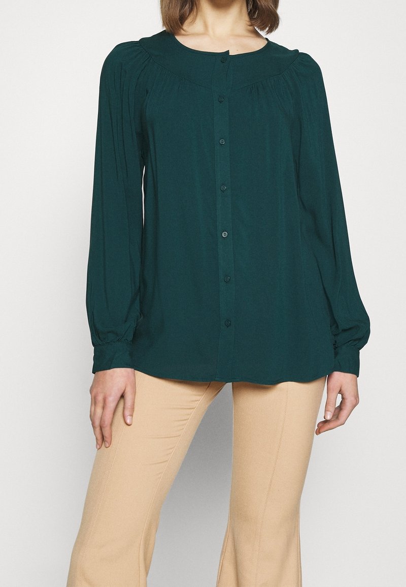 Blouse teal à manches longues avec un col rond, fermeture à boutons, et des détails froncés aux épaules, associée à un pantalon beige ajusté.