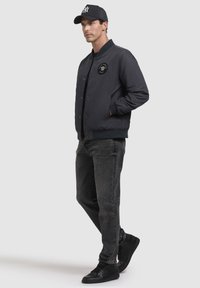 Jachetă bomber de culoare bleu închis, cu o inscripție rotundă, purtată cu blugi negri și adidași negri. Țesătura pare netedă, iar croiala este lejeră.