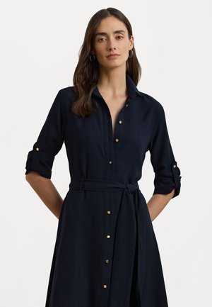Marineblauwe shirtjurk van zacht stof met een knoopsluiting aan de voorkant, gouden knopen en een ceintuur in de taille. Heeft opgerolde mouwen en een kraag.