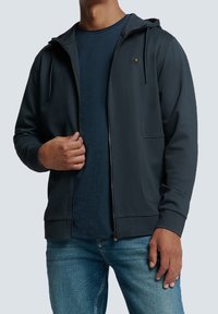 Felpa con zip di un grigio scuro, dotata di tasca frontale, cappuccio regolabile con cordino e un piccolo logo sul petto sinistro, indossata sopra una maglietta blu.