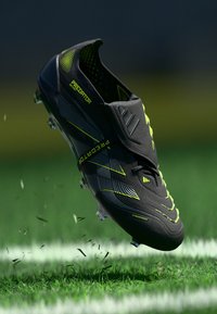 Black soccer cleat with neon green accents, textured surface, and slip-on design. Features a prominent logo and studded outsole for traction.