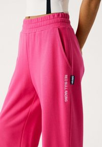 Lyserøde sweatpants lavet af blødt stof, med elastisk talje, sidelommer og "RED BULL RACING" broderet i hvid.