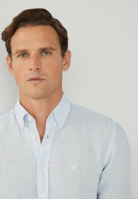 Camicia di lino azzurro chiaro con colletto button-down, emblema ricamato sul petto e trama leggermente trasparente.