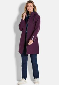 Paarse waterdichte jas met capuchon, voorzien van een rits aan de voorkant, versterkte koordjes, reflecterende accenten en zijzakken, gedragen over donkere jeans en sneakers.