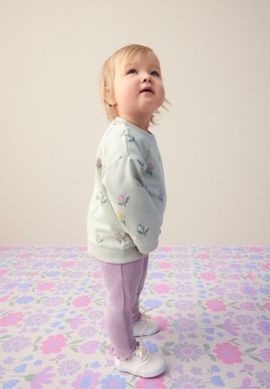 Småbarn står på et pastelblomstret gulvtæppe iført en lyseblå sweatshirt med blomster, lavendelbukser og hvide sneakers, kigger opad.