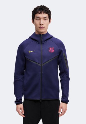 Jeune homme portant une veste zippée bleu marine à capuche, logo jaune sur la poitrine droite et écusson rouge et bleu du FC Barcelone sur la poitrine gauche.