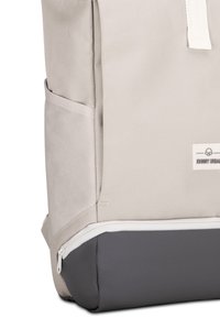 Beige hátizsák textúrált külsejével, szürke aljával és fehér cipzárjával. Oldalsó zsebbel és márka címkével a látható részen.