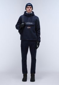 Felpa con cappuccio di velluto blu navale con un patch del logo, abbinata a pantaloni scuri e stivali neri. Gli accessori includono guanti e un berretto abbinato.
