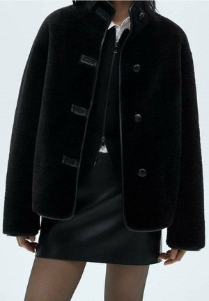 Personne portant une veste noire duveteuse par-dessus un haut noir zippé et une mini-jupe en cuir noire avec des collants noirs transparents.