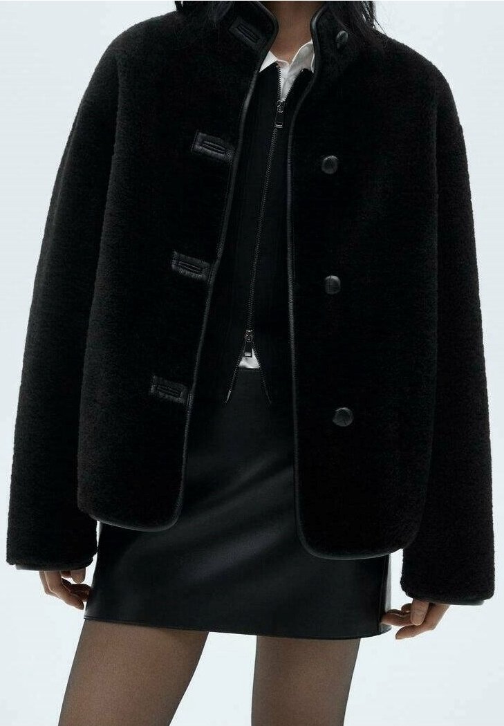 Personne portant une veste noire duveteuse par-dessus un haut noir zippé et une mini-jupe en cuir noire avec des collants noirs transparents.