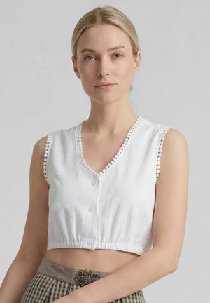 Jonge vrouw met blond haar draagt een wit mouwloos cropped topje met knoopdetails en een geruite rok met een decoratieve riemgesp.