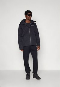 Czarna wodoodporna kurtka z kapturem, zamkiem błyskawicznym i siateczkową podszewką, zestawiona z czarnymi spodniami i butami sportowymi, noszona przez stojącą osobę.