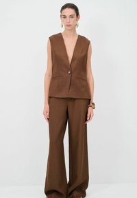 Braun maßgeschneiderte Weste mit tiefem V-Ausschnitt und vorderem Knopf. Kombiniert mit weit geschnittenen braunen Hosen, beide aus glattem Stoff. Minimalistisches Design.